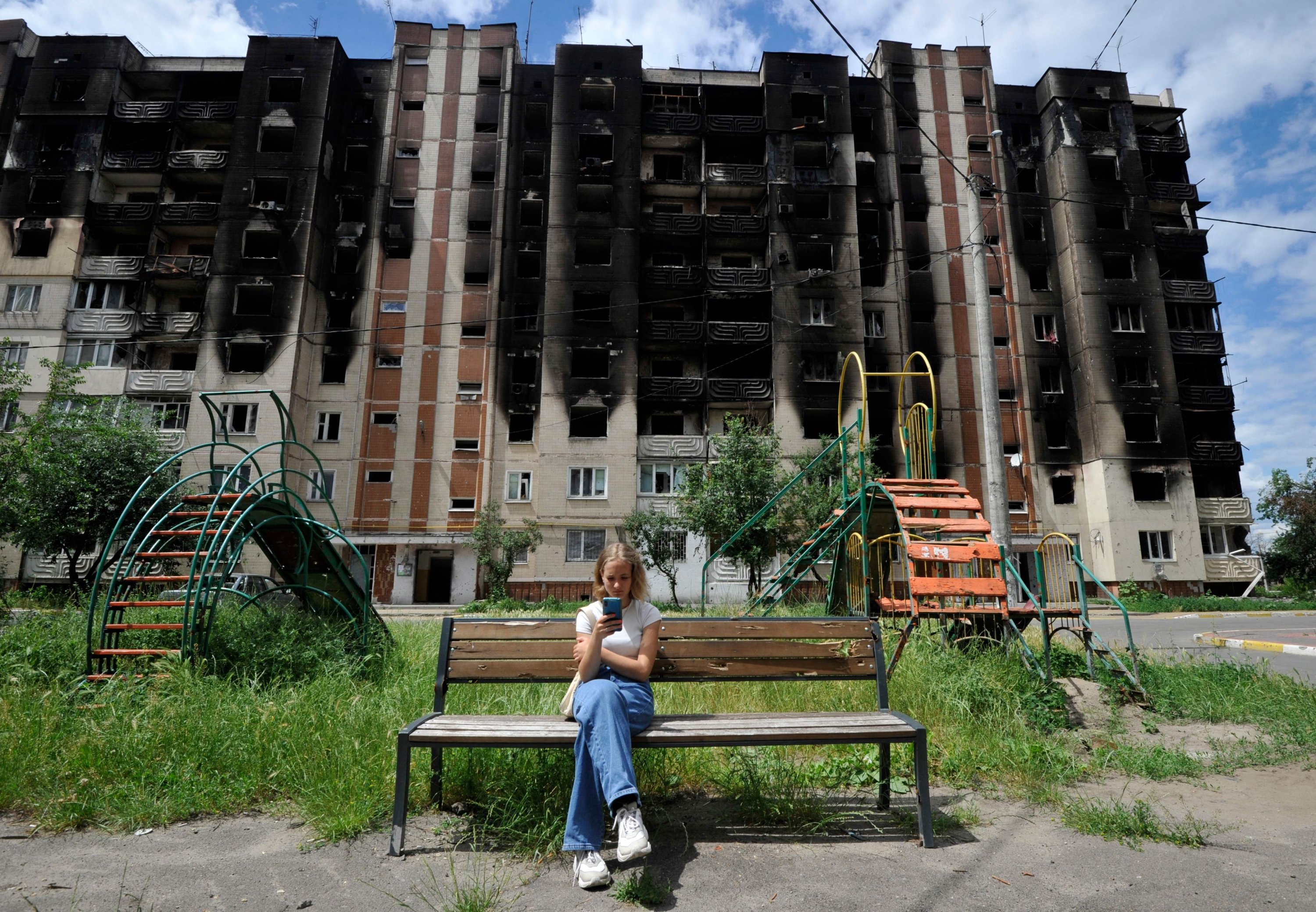 314535 A girl looks at her smartphone as she sits on a bench in front of partially destroyed residential buildings in near Kyiv, Ukraine, June 16, 2022. (AFP Photo)