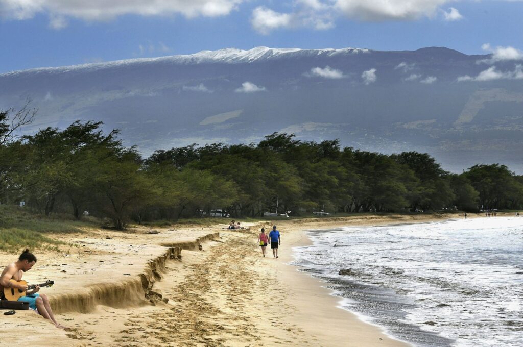 Snow-capped Haleakala serves as a backdrop as Paia, Maui's Scott Picton, (L) plays a guitar and Saskatchewan's Shannon and Dan Runcie walk on Sugar Beach in South Maui, Hawaii, Feb. 11, 2019. (AP Photo)