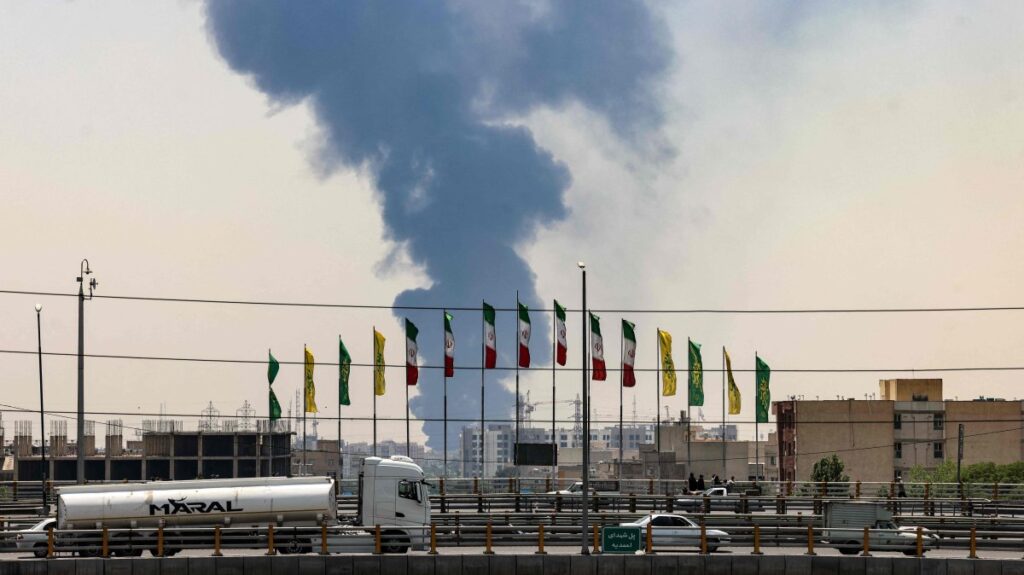 Flags flutter along a bridge as a plume of heavy smoke and fire rise from an oil refinery in southern Tehran, after it was hit in an overnight Israeli strike, June 15, 2025. (AFP Photo)